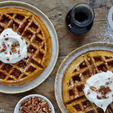 Pumpkin Spice Waffles sprinkled with powdered sugar topped with maple syrup, whipped cream and chopped pecans