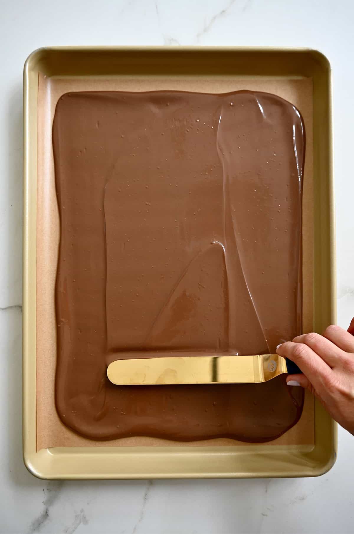 A hand holding an offset spatula spreads melted chocolate atop a parchment paper-lined baking sheet to make chocolate bark.