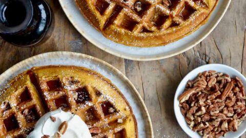 Two Pumpkin Spice Waffles dusted with confectioners' sugar topped with maple syrup, whipped cream and chopped pecans