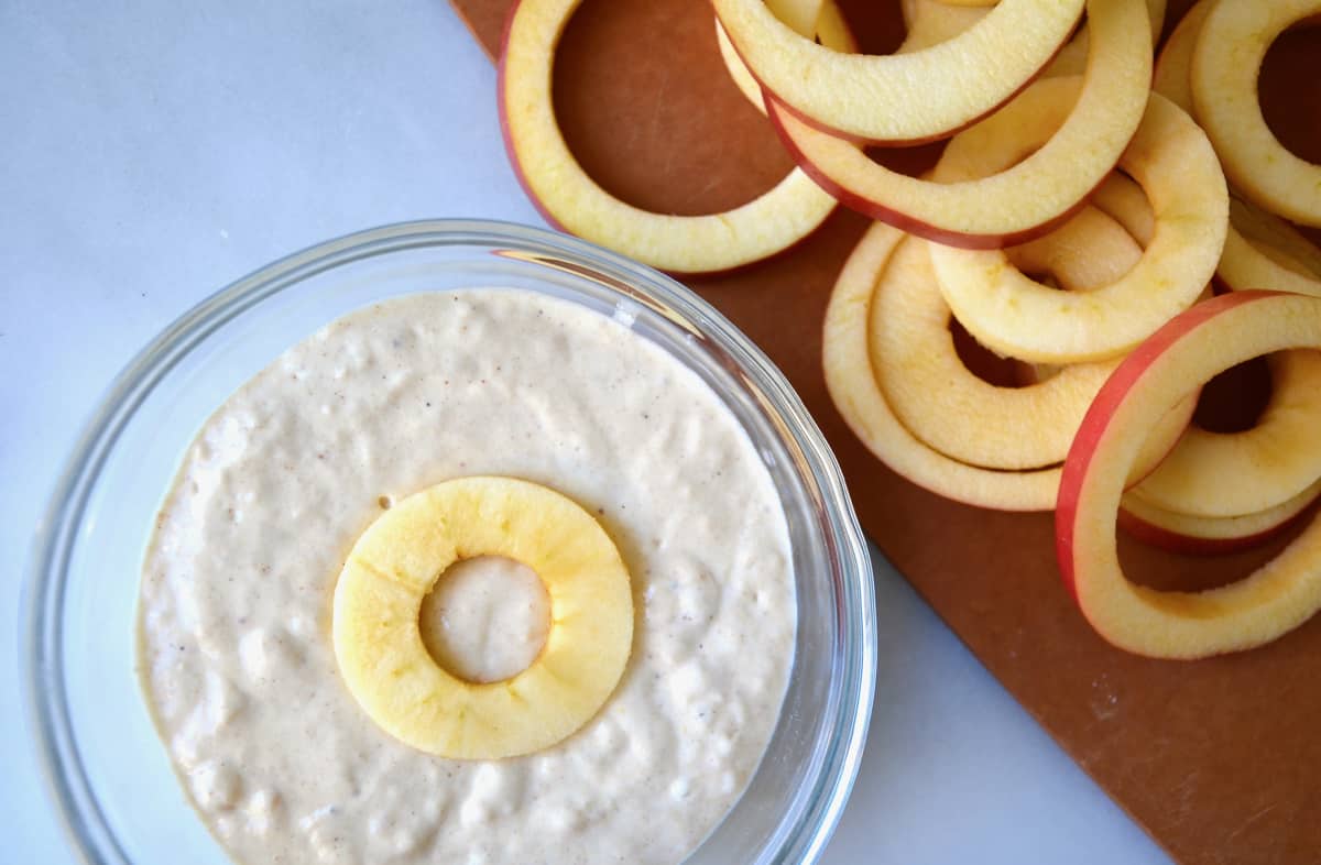 An apple ring sits atop batter in a clear bowl. More apple rings on a cutting board are next to the bowl.