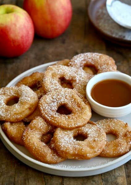 Golden brown apple fritter rings dusted with powdered sugar on a white platter with caramel sauce for dipping and fresh apples in the background.