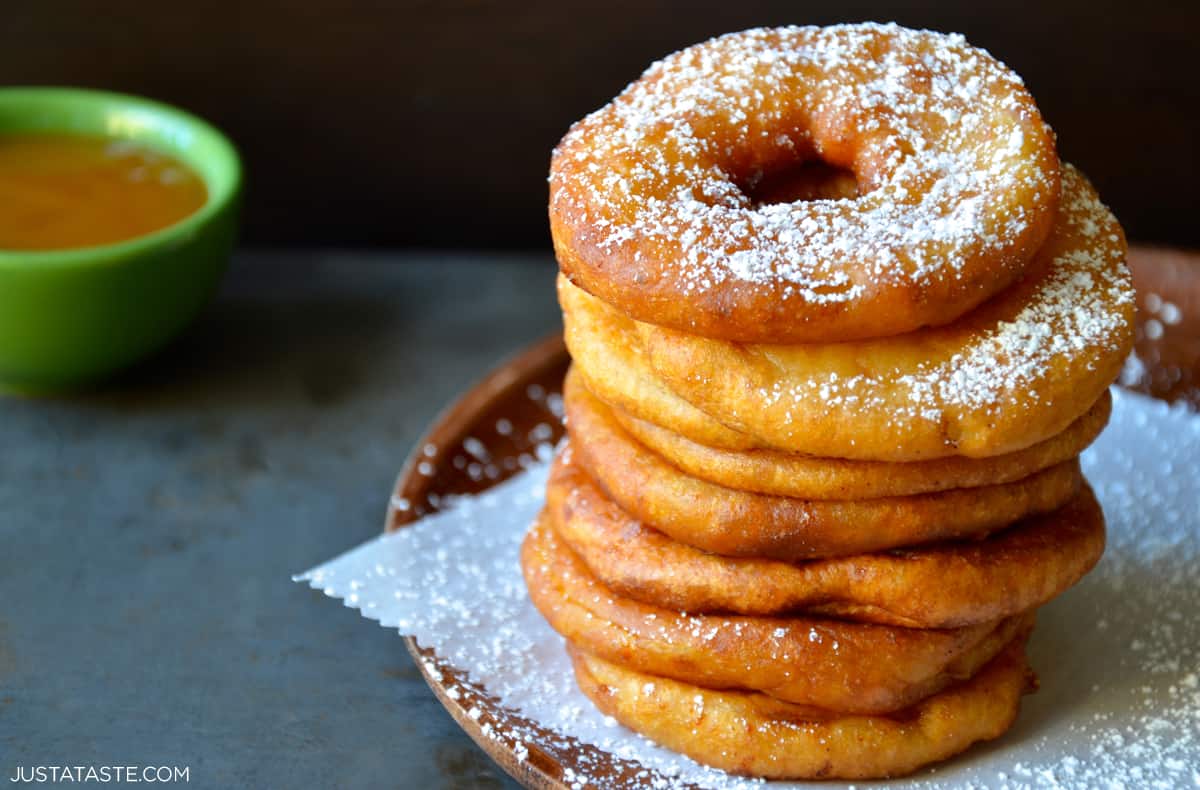 A stack of apple fritter rings dusted with powdered sugar atop a sheet of parchment paper on a plate.