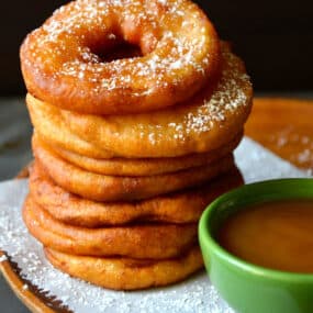 A tall stack of deep fried apple fritter rings next to a small green bowl filled with caramel sauce.