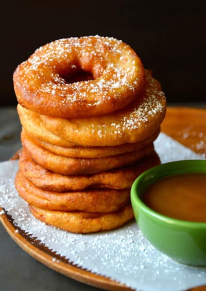 A tall stack of deep fried apple fritter rings next to a small green bowl filled with caramel sauce.