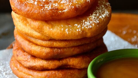 A tall stack of deep fried apple fritter rings next to a small green bowl filled with caramel sauce.