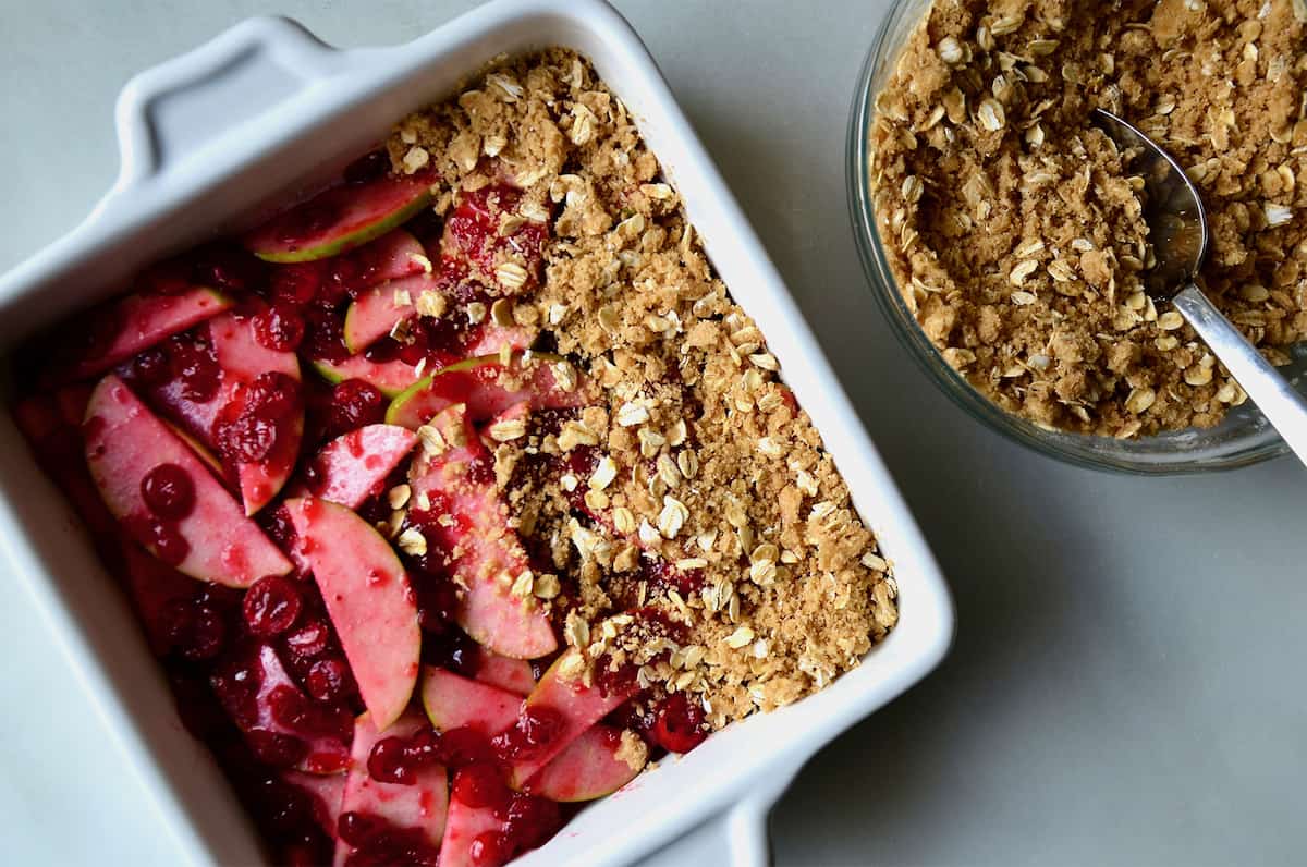 A square baking dish containing apple cranberry crisp next to a clear bowl containing crisp topping.