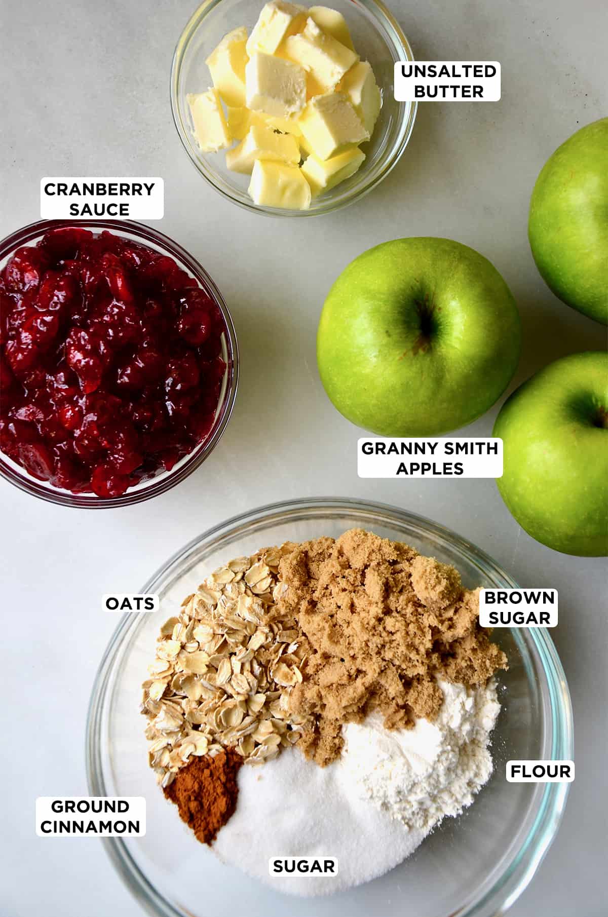 Granny Smith apples next to a bowl containing cranberry sauce, a bowl with cubed butter, and a bowl containing oats, brown sugar, flour and spices.