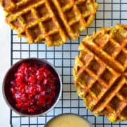 A top-down view of two Leftover Stuffing Waffles atop a wire cooling rack next to small bowls containing cranberry sauce and gravy.