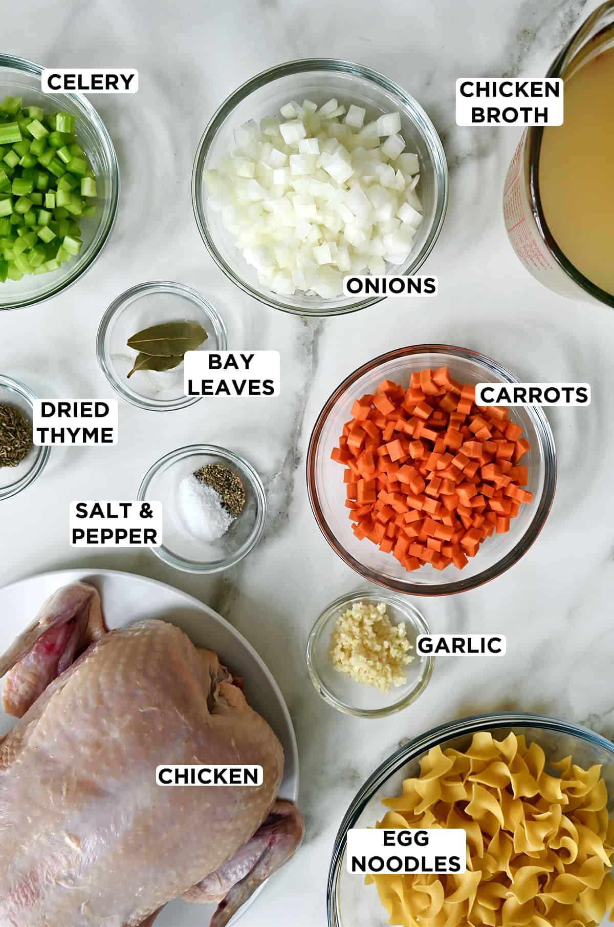 Various sizes of clear bowls containing diced celery, onions, carrots, bay leaves, minced garlic, dried thyme, egg noodles, chicken broth and a whole chicken.