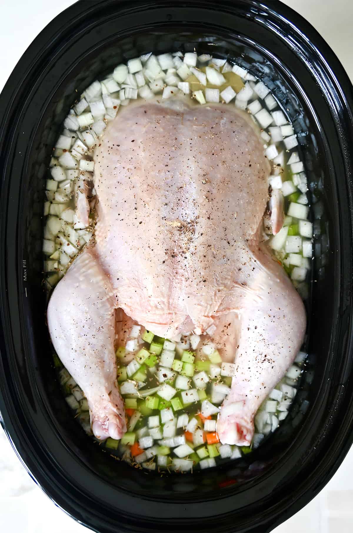 A whole roasting chicken in a slow cooker with diced veggies and broth.