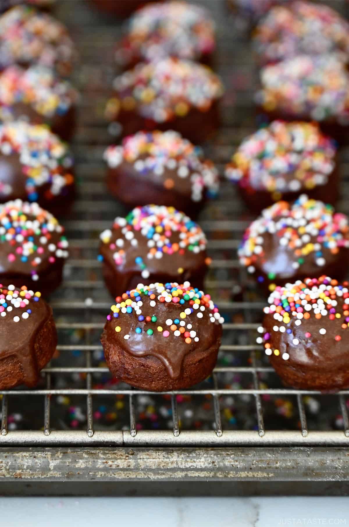 Glazed chocolate doughnut holes with rainbow sprinkles on a wire cooling rack.