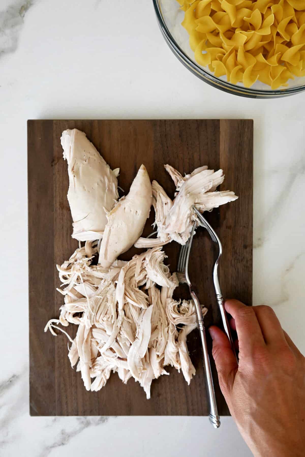A woman's hand reaches for one of two forks on a cutting board with shredded chicken.