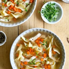 Two bowls containing chicken noodle soup next to a small bowl containing micro greens.