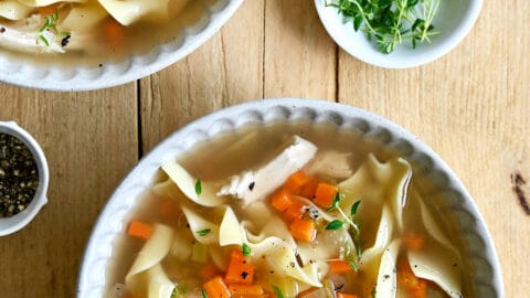 Two bowls containing chicken noodle soup next to a small bowl containing micro greens.