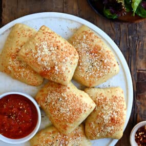 A top-down view of Cheesy Chicken Pizza Pockets on a large white plate next to a small bowl containing pizza sauce