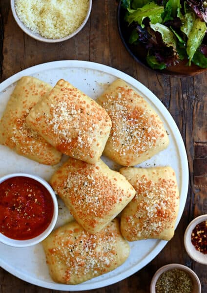 A top-down view of Cheesy Chicken Pizza Pockets on a large white plate next to a small bowl containing pizza sauce