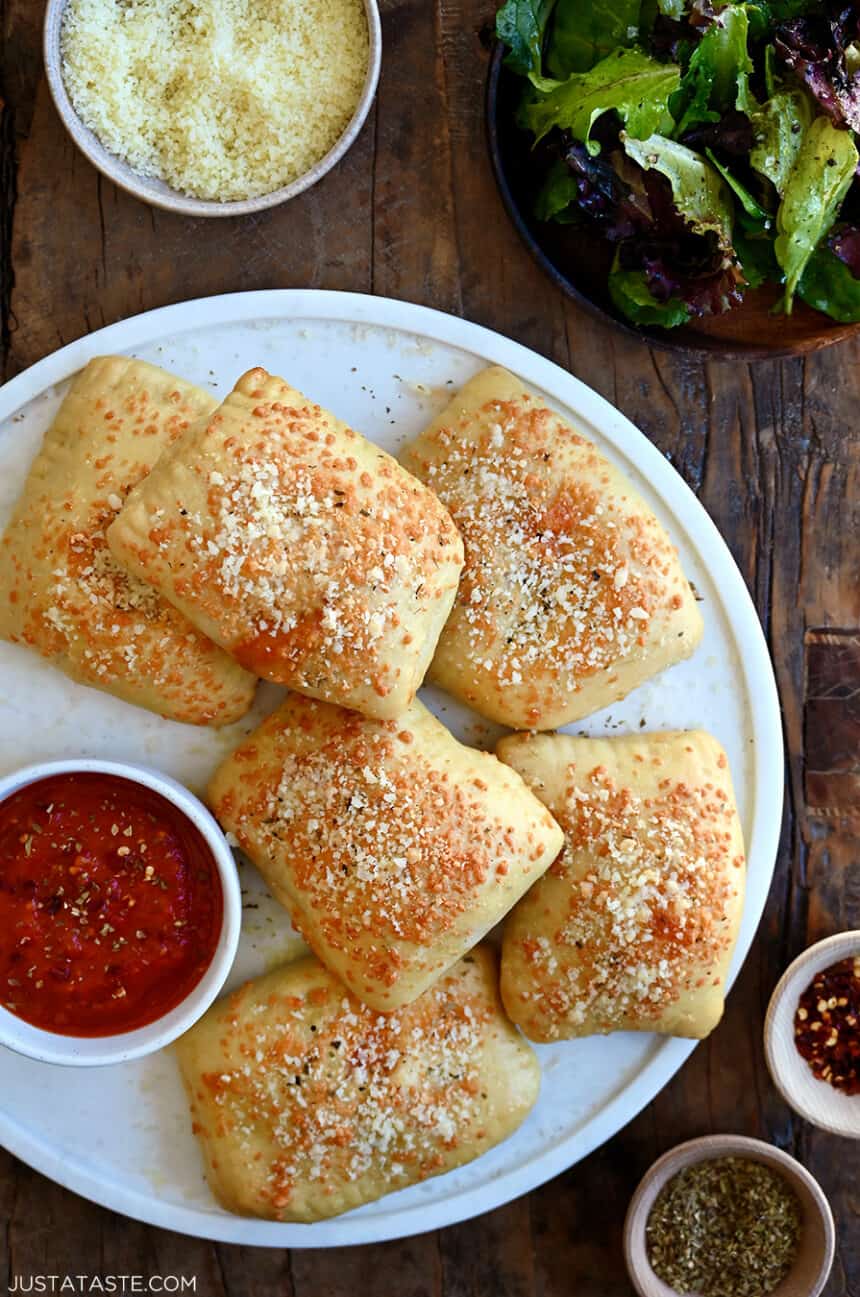 A top-down view of Cheesy Chicken Pizza Pockets on a large white plate next to a small bowl containing pizza sauce
