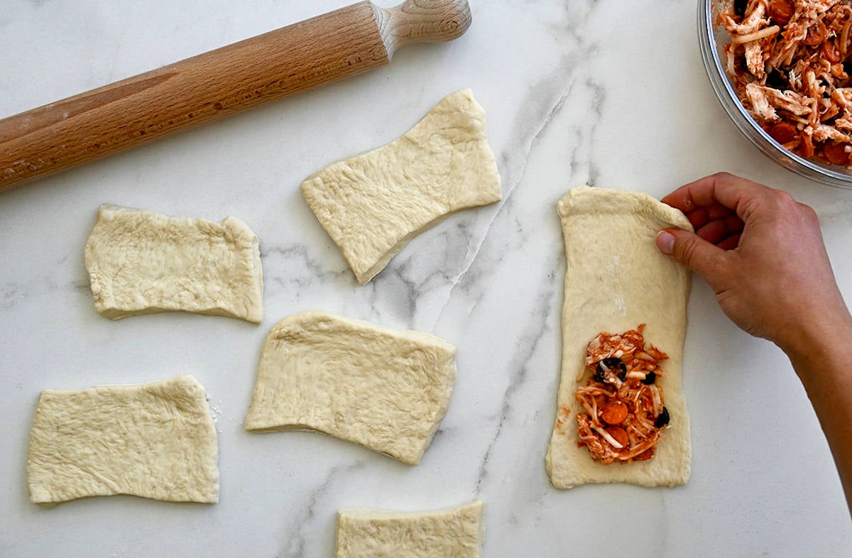 A rolling pin next to pieces of rectangular pizza dough with one piece topped with mini pepperoni, cheese and black olives.