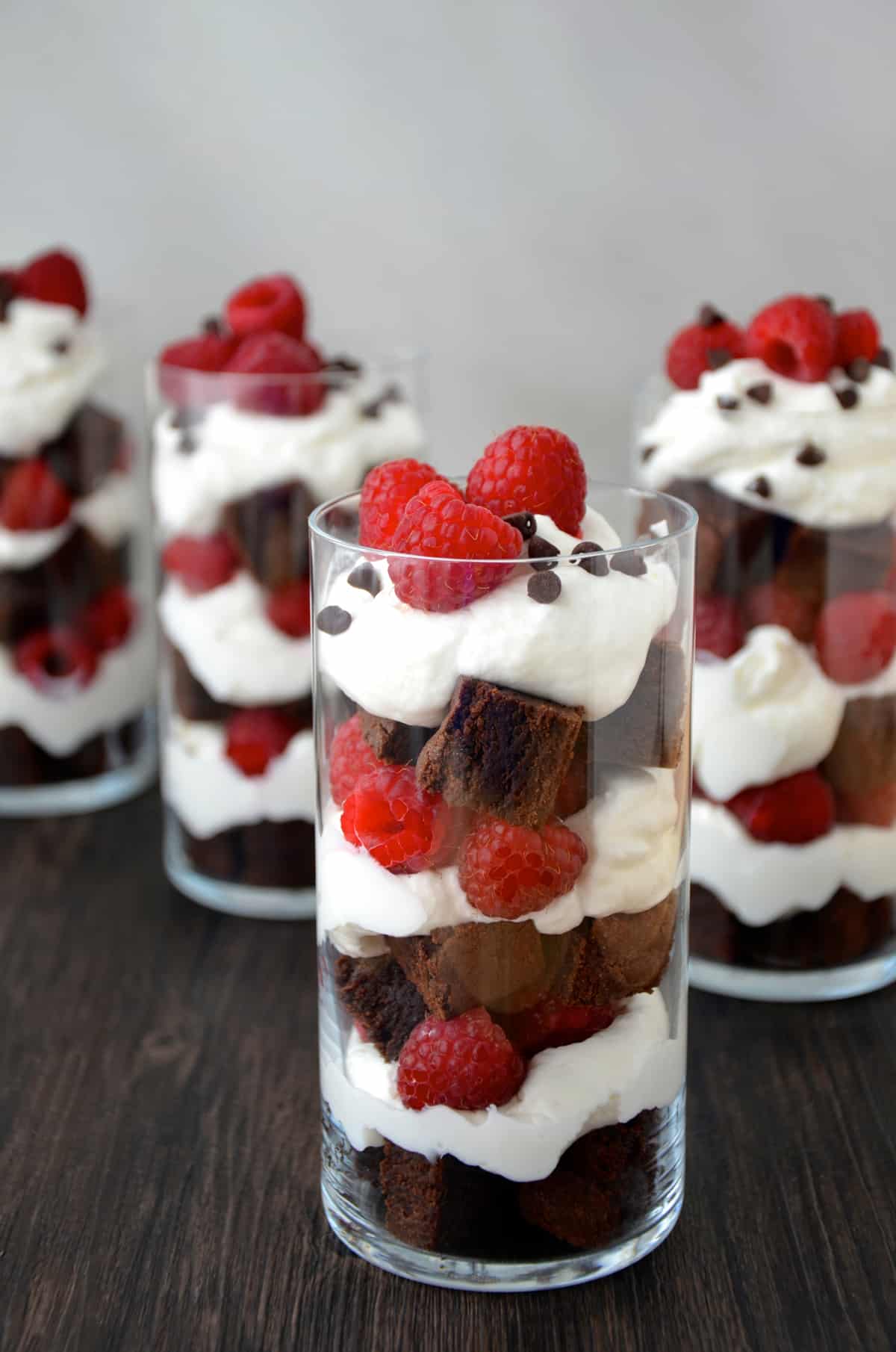 Several parfait glasses filled with layers of brownie pieces, whipped cream and fresh raspberries.