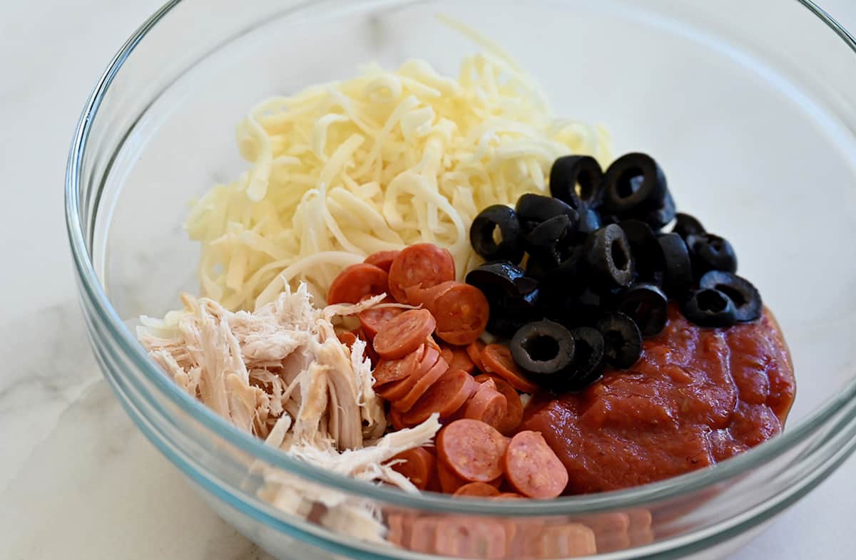 A clear bowl containing shredded chicken, mini pepperonis, black olives, shredded cheese and marinara sauce.