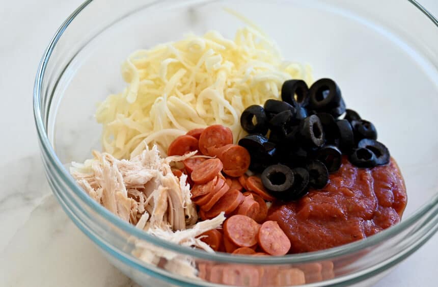 A clear bowl containing shredded chicken, mini pepperonis, black olives, shredded cheese and marinara sauce