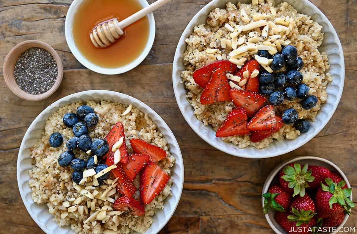 Two bowls with creamy coconut quinoa topped with fresh berries, slivered almonds, and chia seeds next to small bowls containing strawberries, honey and chia seeds.