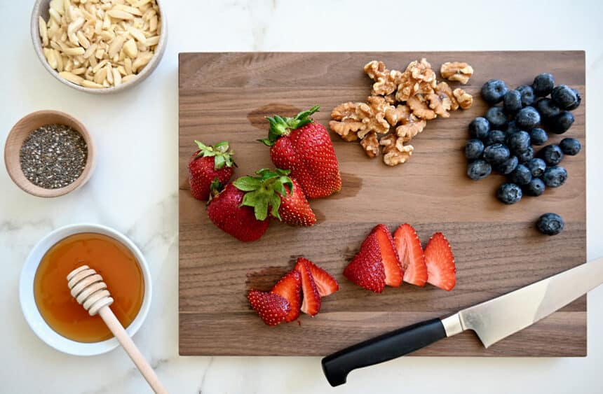 A top-down view of sliced strawberries, walnuts, fresh blueberries, and a sharp knife atop a cutting board next to small dishes containing honey, chia seeds and chopped nuts