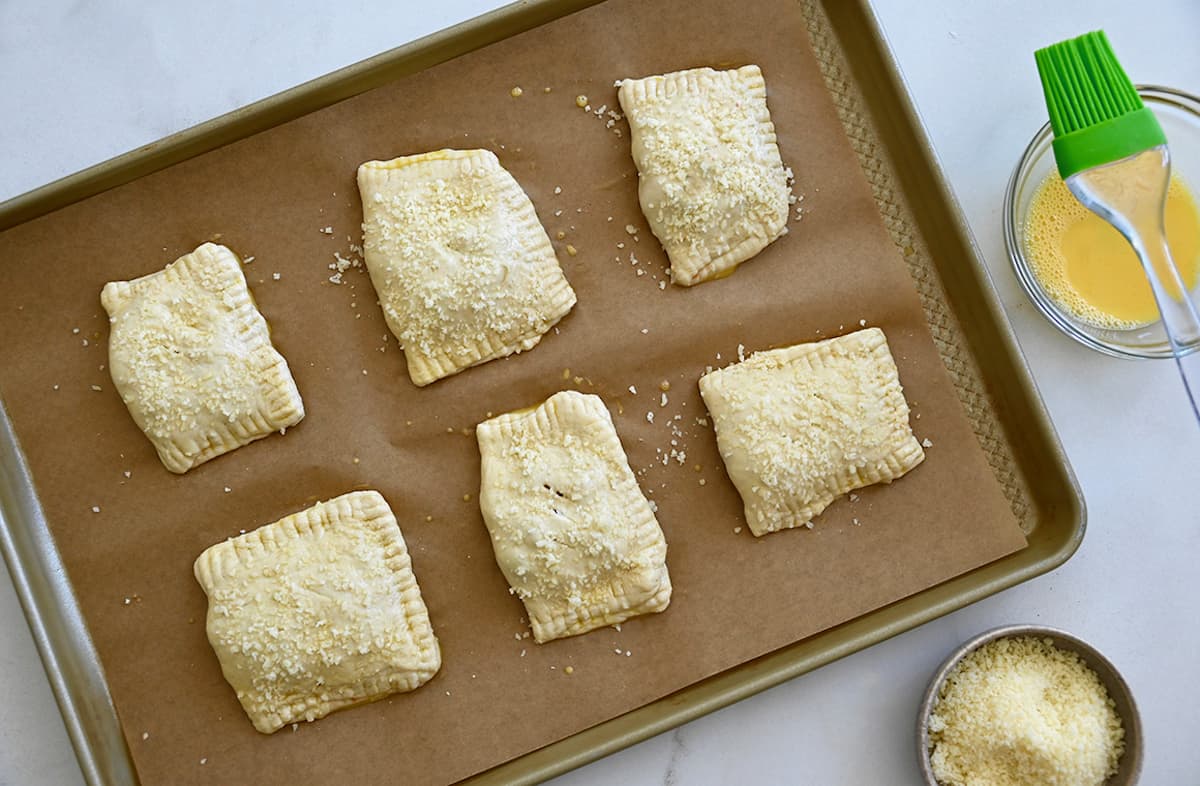 Unbaked homemade pizza pockets brushed with an egg wash and sprinkled with parmesan cheese on a baking sheet lined with parchment paper.