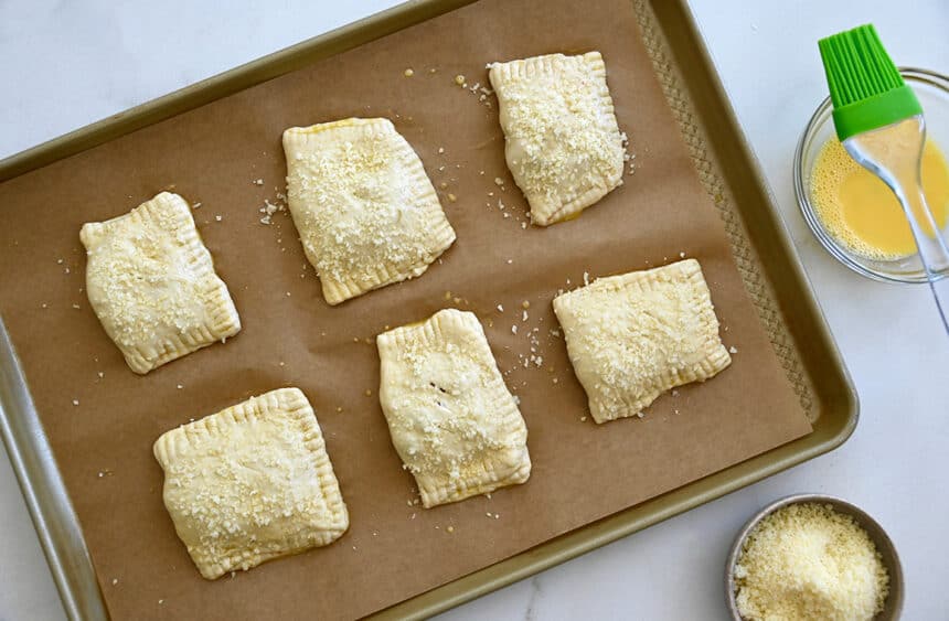 A top-down view of unbaked pizza pockets on a parchment paper-lined baking sheet next to a small bowl containing an egg wash