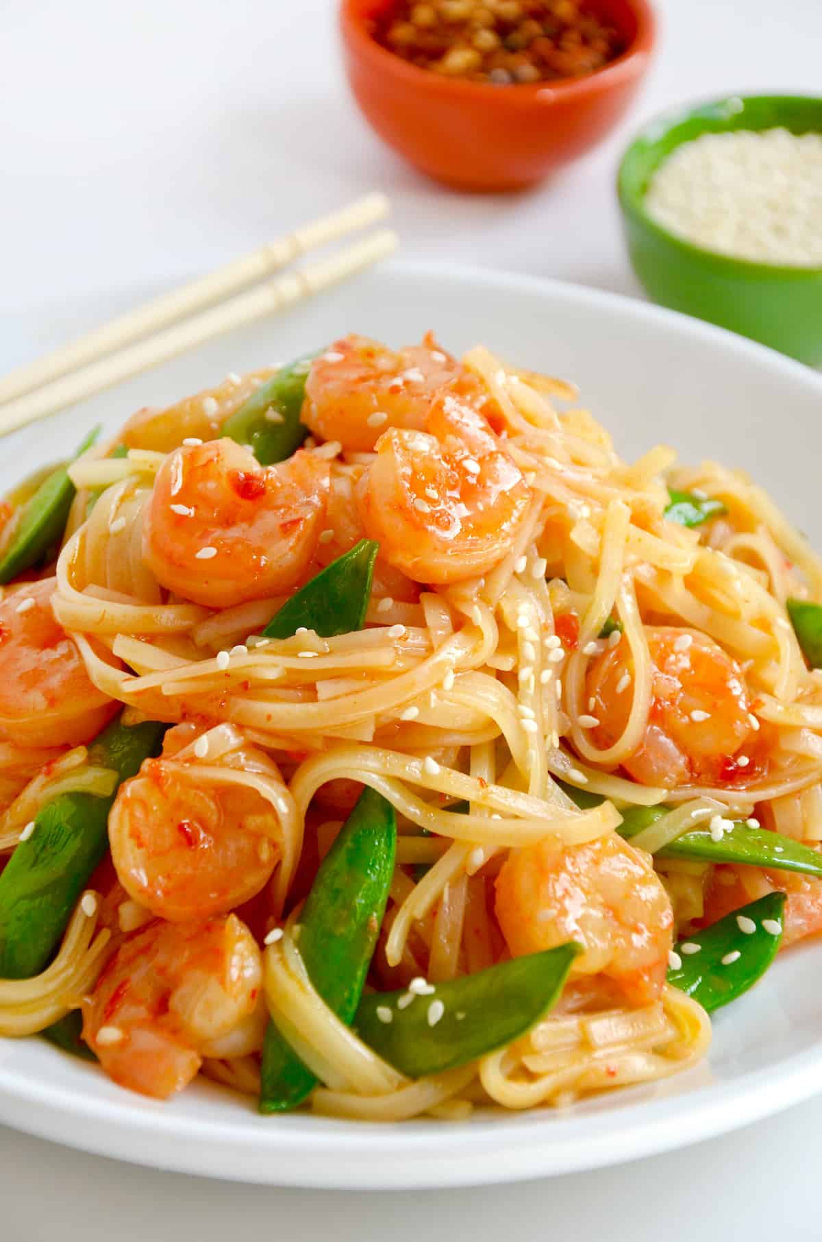 Stir-fried shrimp with snap peas and sesame seeds atop noodles in a white bowl with chopsticks.