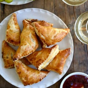 A top-down view of Baked Brie Bites with Jam piled on a plate next to a small bowl containing jam and two glasses of white wine