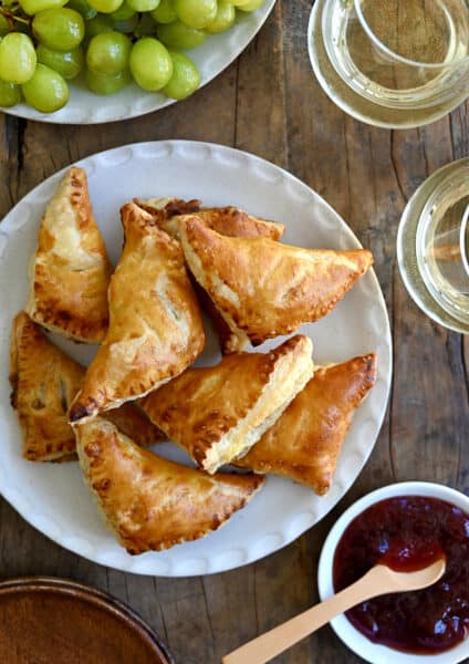 A top-down view of Baked Brie Bites with Jam piled on a plate next to a small bowl containing jam and two glasses of white wine