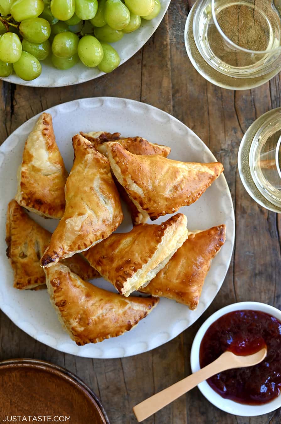 A top-down view of Baked Brie Bites with Jam piled on a plate next to a small bowl containing jam and two glasses of white wine