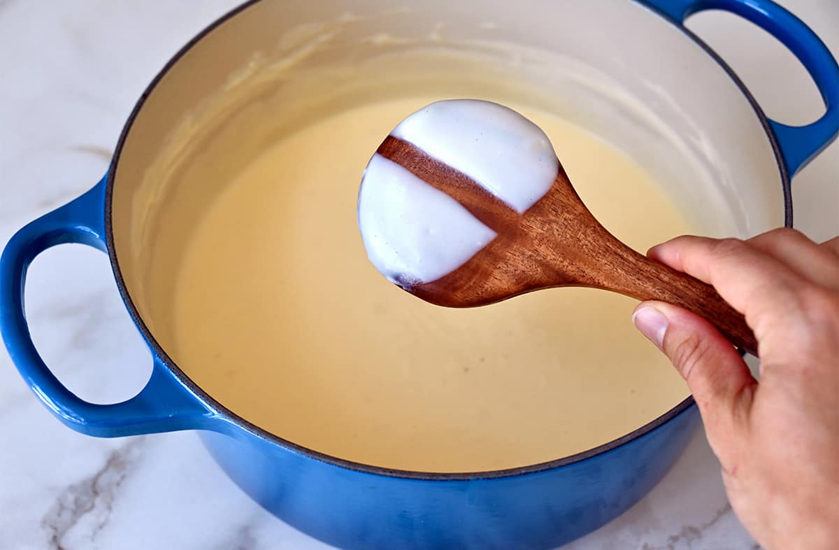 Thick cheese sauce on the back of a wooden spoon over a large blue stockpot.