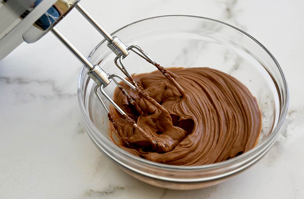 A handheld mixer resting in a clear bowl that contains a whipped Nutella-cream cheese mixture