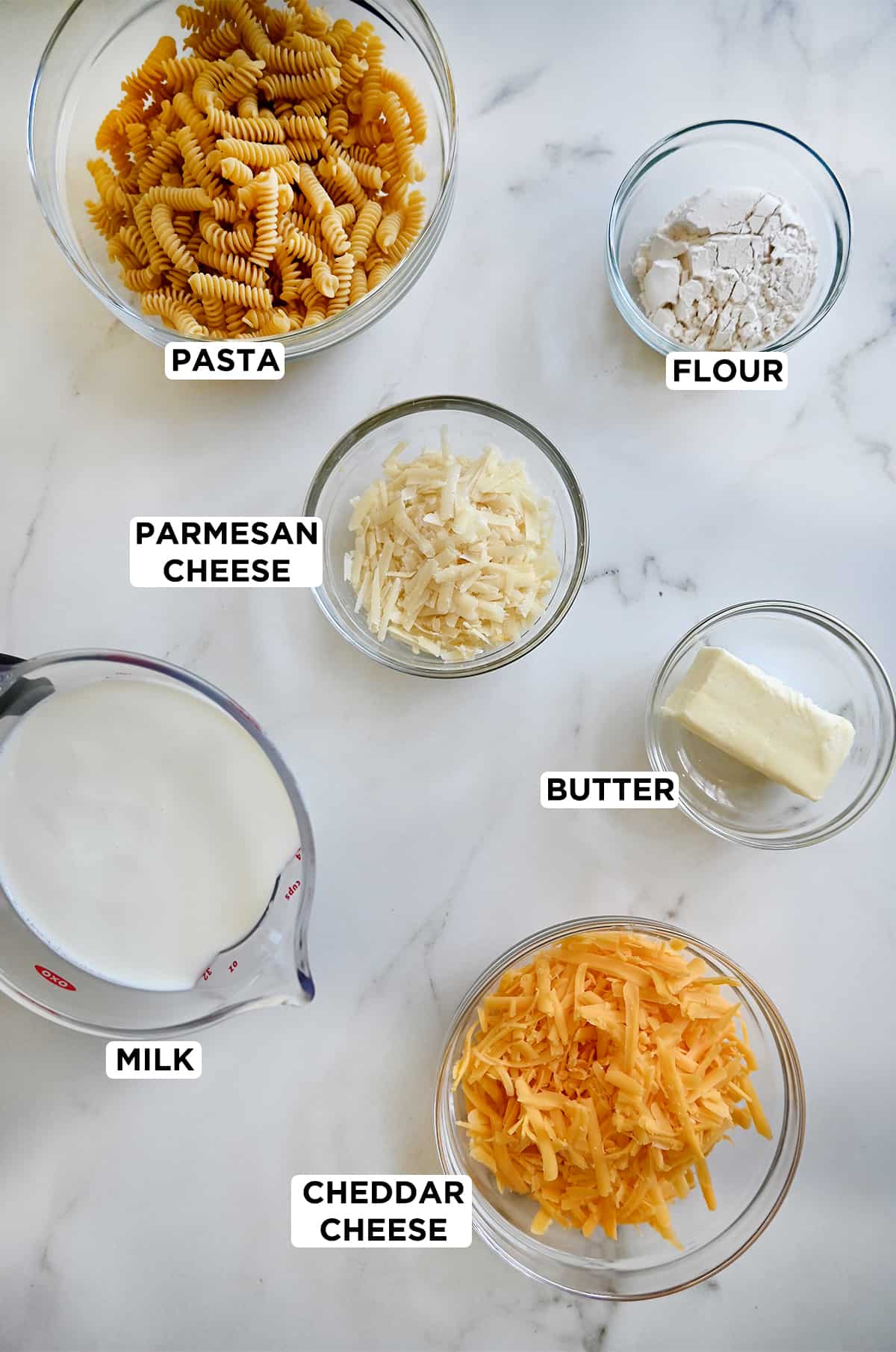 Various sizes of clear bowls containing the ingredients needed to make mac and cheese, including uncooked pasta, flour, Parmesan cheese, butter, cheddar cheese and milk.