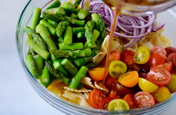 Dressing being poured over cherry tomatoes and asparagus in clear bowl