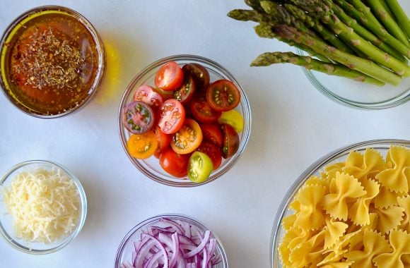 Various sizes of clear bowls containing italian dressing, halved cherry tomatoes, asparagus, bow tie pasta, sliced red onion and parmesan cheese