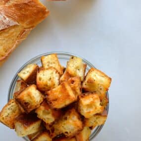 Homemade croutons in a small glass bowl next to a baguette.