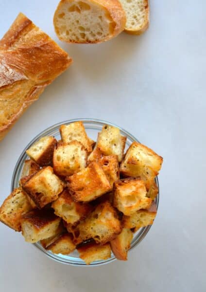 Homemade croutons in a small glass bowl next to a baguette.