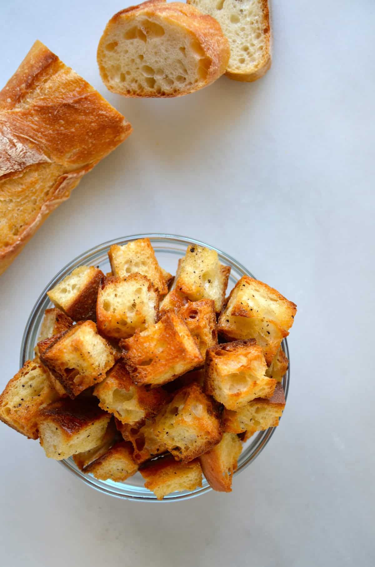 Homemade croutons in a small glass bowl next to a baguette.