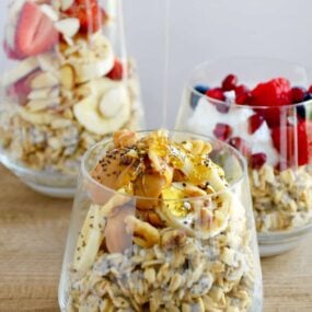 A clear glass containing oats topped with bananas, peanut butter and chia seeds with honey being drizzled on top