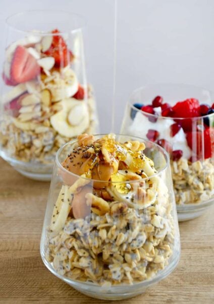 A clear glass containing oats topped with bananas, peanut butter and chia seeds with honey being drizzled on top