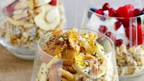 A clear glass containing oats topped with bananas, peanut butter and chia seeds with honey being drizzled on top