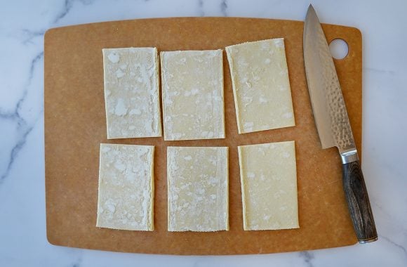 Cutting board containing six rectangles of cut puff pastry next to a sharp knife 