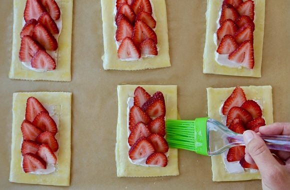 Hand with brush brushing egg wash on pastry with strawberries 