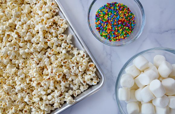 Baking sheet with popcorn next to a small bowl with sprinkles and a large bowl with marshmallows 