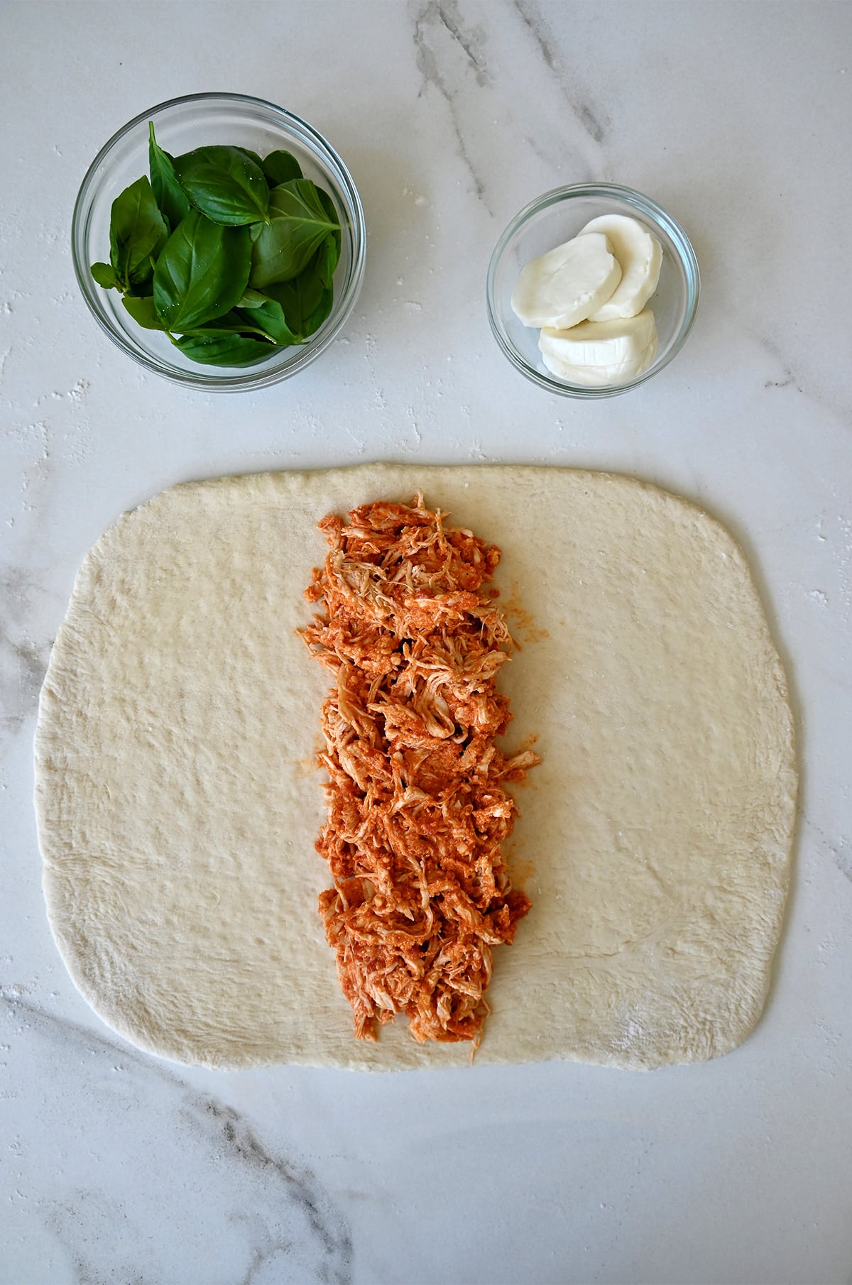 Chicken filling atop pizza dough next to small bowls containing fresh mozzarella cheese and fresh basil leaves.