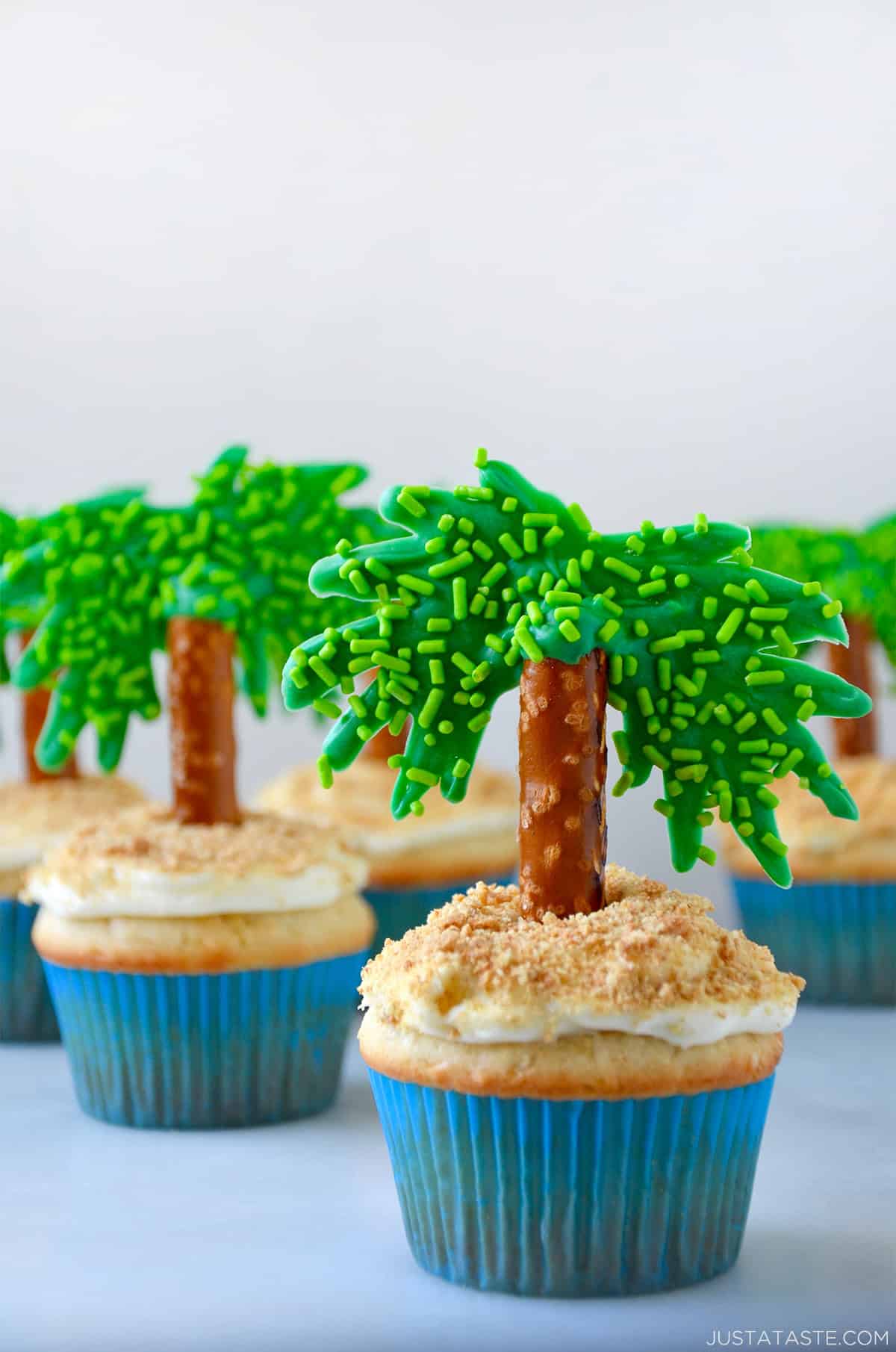 Moist coconut cupcakes topped with cream cheese frosting, crushed graham crackers and pretzel palm tree toppers to look like mini tropical islands.