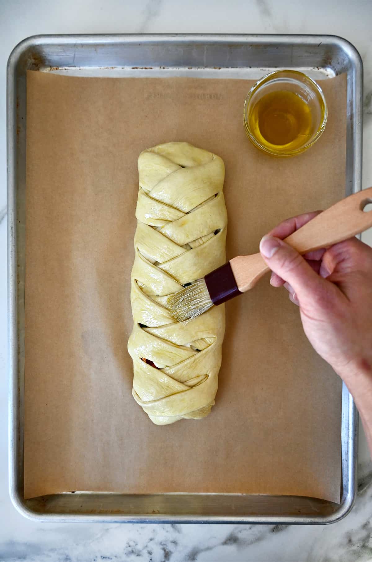 A hand holding a pastry brush applies olive oil to a braided pizza bread on a parchment paper-lined baking sheet.